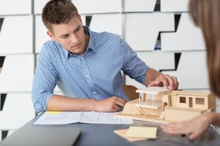 Young Male Designer Assembling Wooden Pieces into a House Miniature on Top of the Table with Blueprint.
